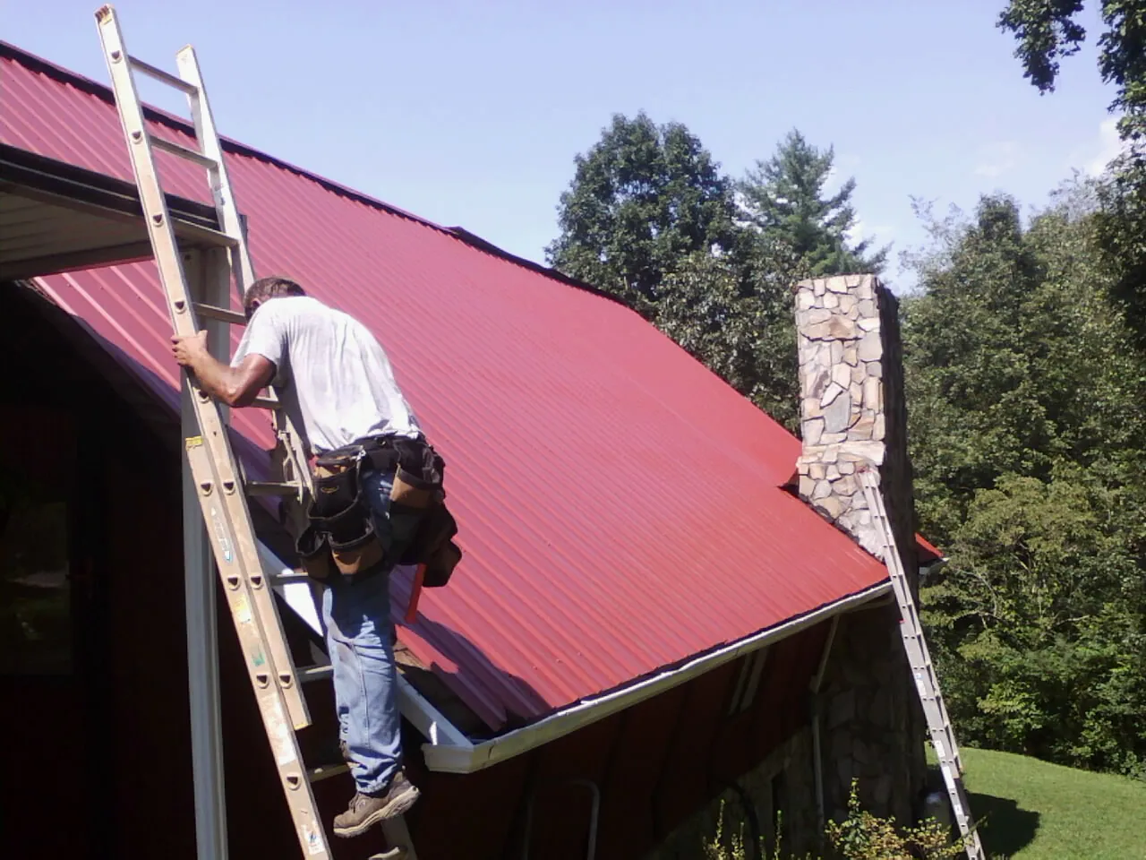 Roofer on a ladder beside a red metal roof and stone chimney — Soffit & Fascia Repair by Roofing Brothers in Valdese